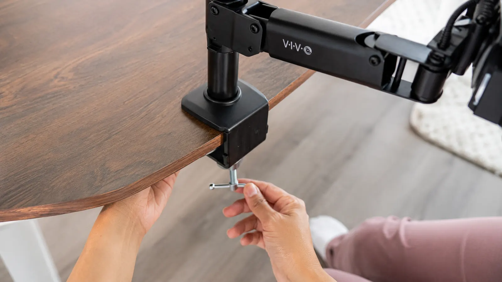 Close-up of hands tightening the clamp bolt on a VIVO monitor arm mounted to a desk edge